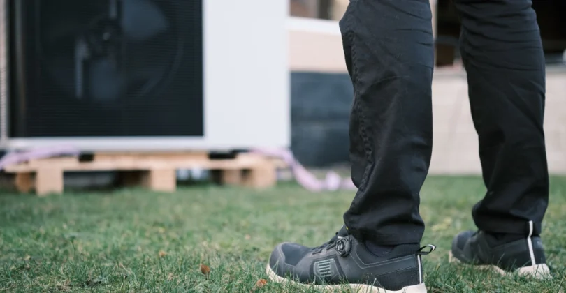 Legs of installer standing on grass in front of an outdoor unit/heat pump.