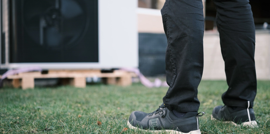 Legs of installer standing on grass in front of an outdoor unit/heat pump.